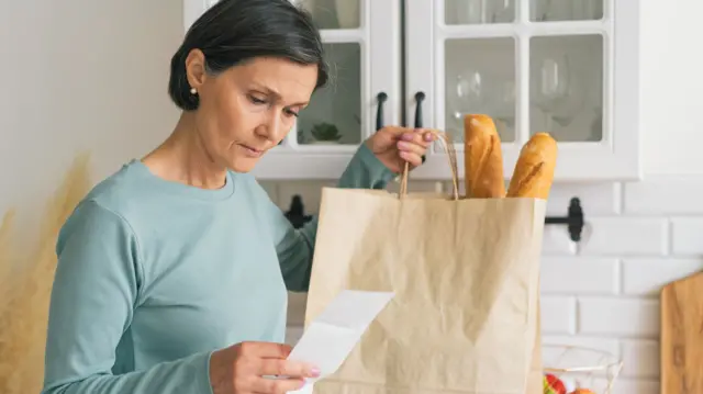 Woman looking at receipt from grocery shop, while holding paper bag containing baguettes