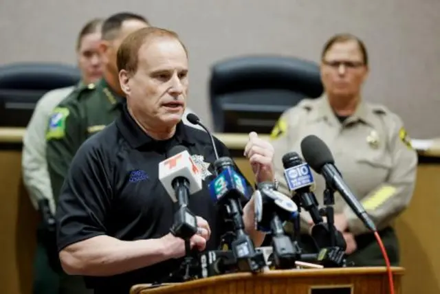 A man in a dark polo shirt stands behind a bank of microphones
