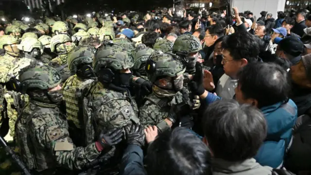 A group of soldiers in combat uniform and helmets are confronted by civilians