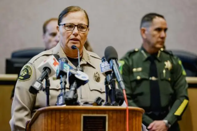 A woman in glasses and a sheriff's uniform stands at a bank of microphonesR