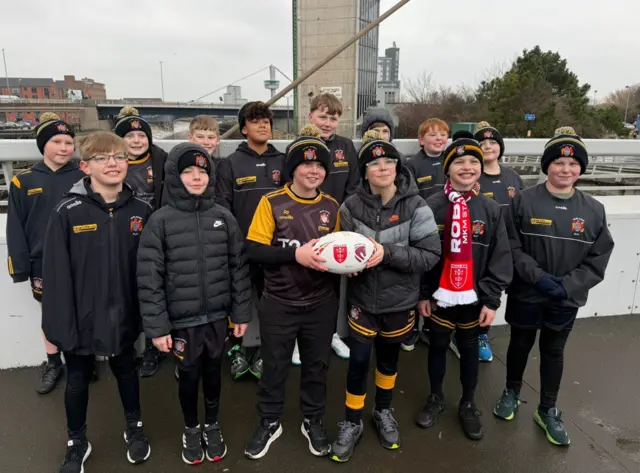 A group of young boys wearing the same black and yellow sports outfits and woolly hats with a yellow bobble on them. They are holding a rugby ball and standing on a bridge