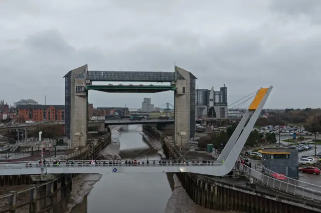 A landscape photograph of the River Hull bridge with several spectators dotted across it. A car park can be seen on the right side of the river and industrial buildings on the left and in the distance.