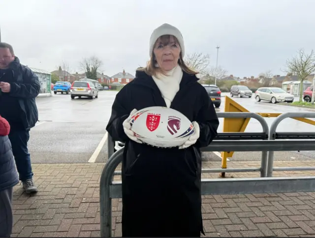 A woman in a white beanie and shoulder-length brown hair is wearing a black coat, white gloves and a scarf and holding a rugby ball in front of a car park.