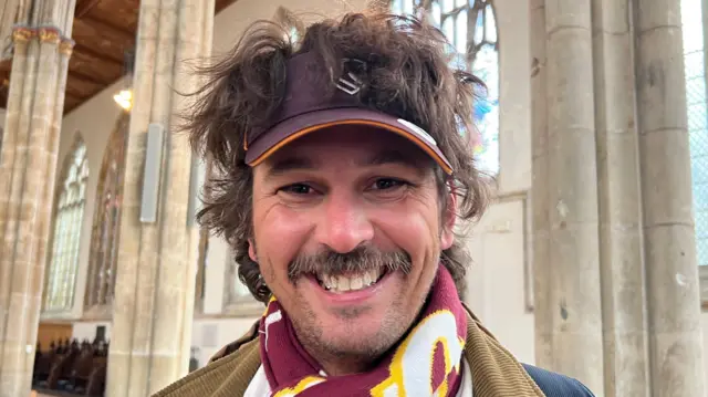 The picture shows a man smiling at the camera. He has brown hair and is wearing a purple cap. He's wearing a burgundy, white and yellow patterned scarf. He is standing in a church, and there are multiple stone pillars behind him.