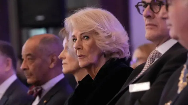 Queen Camilla sat in a row of people at a concert. She is wearing a navy coat, she is sat beside a man wearing glasses.
