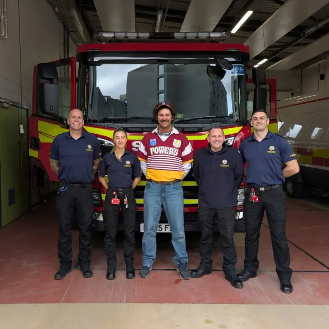 Five people, including one women and four men, are stood in front of a red and yellow fire truck. The man in the middle is wearing jeans, a cap and a shirt that says 'Powers' on it. The other people are wearing navy shirts and black trousers, all with their hands behind their backs.