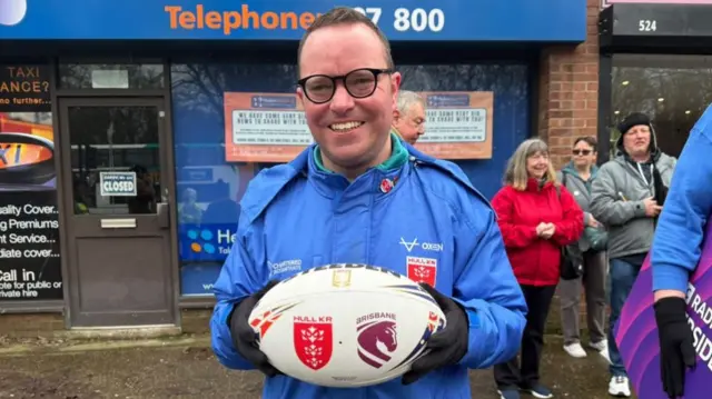 A man with short brown hair is wearing a blue coat and smiling as he holds a rugby ball. A shop can be seen behind him with other people stood around.
