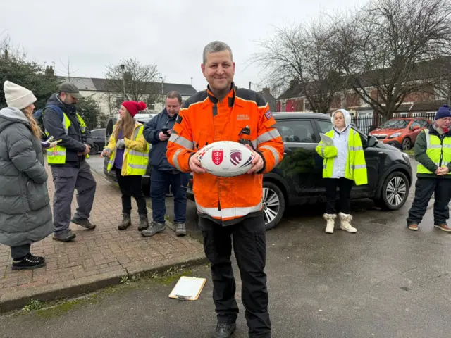 A man in an orange high vis coat, black trousers, grey hair and holding a rugby ball is smiling at the camera. He is stood in a car park with several people behind him wearing yellow hi vis.
