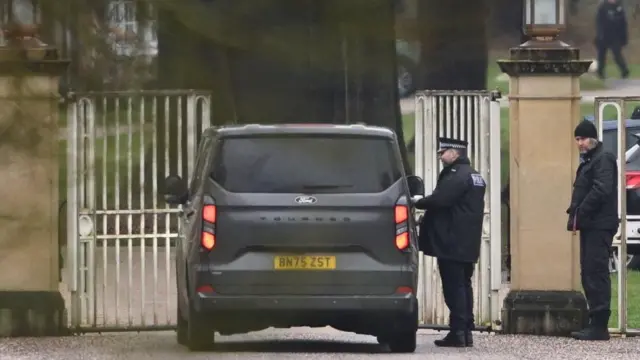 A police officer stands next to a vehicle outside Royal Lodge as a black vehicle arrives at the gate