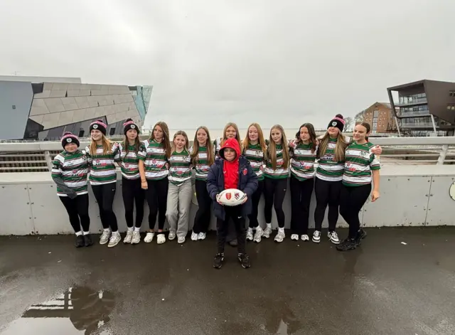 A group of girls in green and white striped tops with a young person stood at the front of them with a red hoodie and navy jacket holding a rugby ball.