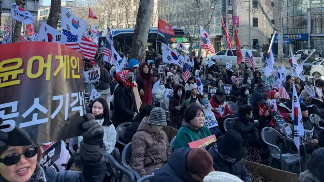 A crowd gathers on a road sitting on chairs and holding the South Korean flag