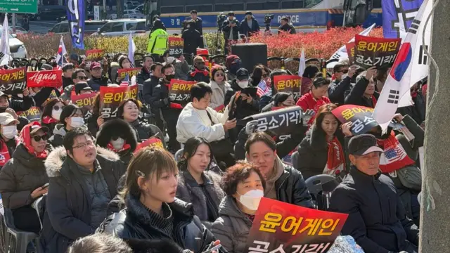 A crowd holding pro-Yoon banners sitting down on a road near the court