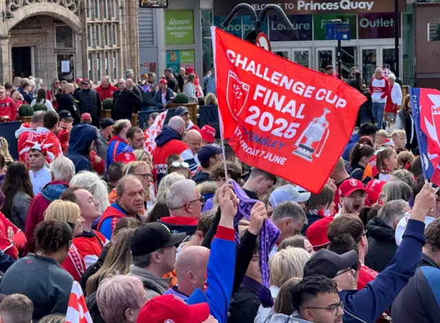 A crowd of rugby fans wearing caps and holding up flags, one large one at the front reads 'Challenge Cup Final 2025'. They are stood in what appears to be a market square.