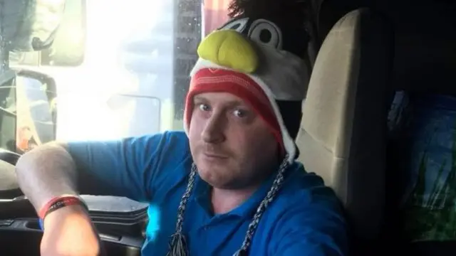 James Barlow pictured inside the cab of a lorry wearing a novelty Hull FChat. He is wearing a blue polo shirt and a watch on his left wrist.