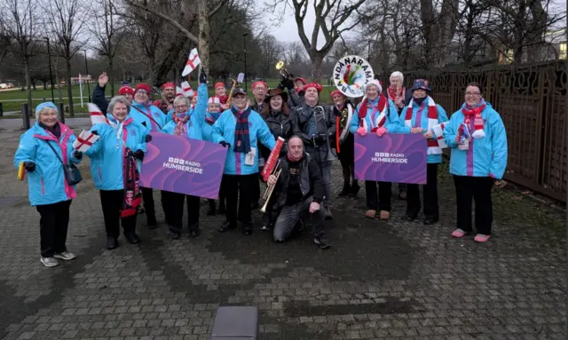 A brass band and smiling crowd wearring turquoise coats