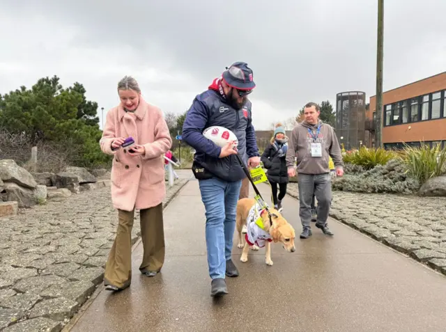 A woman with a pink coat is walking along a path next to a man carrying a rugby ball and looking down at his guide dog, a yellow labrador. Two other people, a man and woman, can be seen walking next to him.
