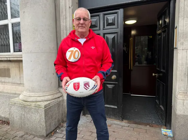 A man with short grey hair is wearing thin rectangular glasses and holding a rugby ball. He has a red jacket on, blue jeans and a large 70 birthday badge on his chest.