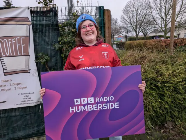 Maddie Tyrrell holding a large BBC Radio Humberside, red top and blue bobble hat. She is smiling at the camera
