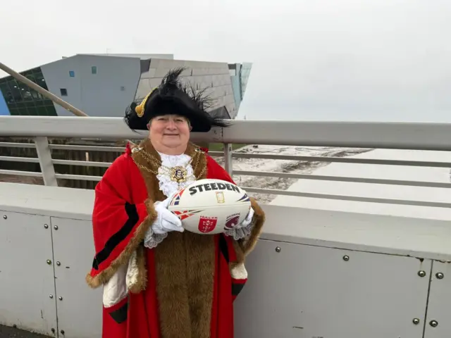 A mayor is dressed up in uniform with a red gown and black traditional hat and is smiling as she holds a rugby ball on a bridge, with a river in the background and modern-looking building.