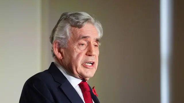 Gordon Brown speaking at an event, wearing a dark blue suit. He is in the middle of speaking and gesticulating slightly with his arms bent at the elbow.