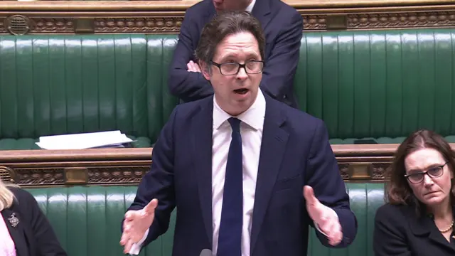 Alex Burghart wears a navy suit and tie as he speaks in the House of Commons