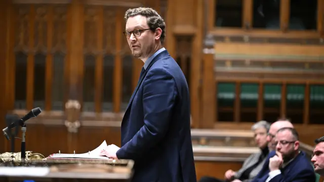 Jones wears a suit and glasses while thumbing through a binder at a podium in the House of Commons on Mondays, with MPs behind him.