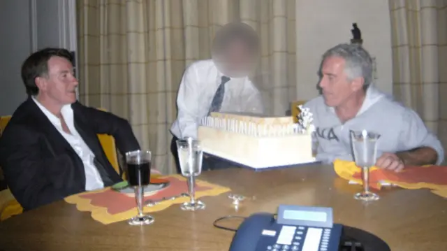 Mandelson sits at a table and looks at a birthday cake that is being held by a person, whose face is blurred. Epstein sits across from him and appears to be blowing out candles.