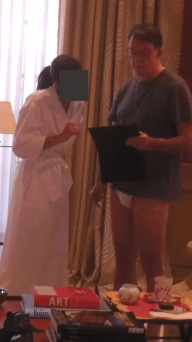 Peter Mandelson is seen standing in a room in his underwear, while a female stands beside him in a white robe. Her face is redacted.