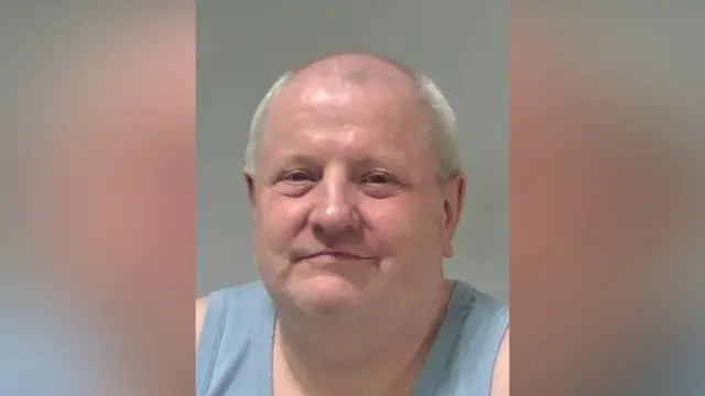 Steve Wright's custody photo. He is smiling at the camera and is wearing a light blue vest top. He has is bald on the top of his head, with shaved white hair around the sides.