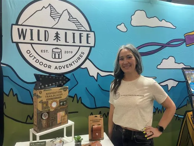 Jenelle Peterson stnads in front of a sign that has a mountain printed on it, she is wearing a white shirt, with her hand on her hip and is smiling