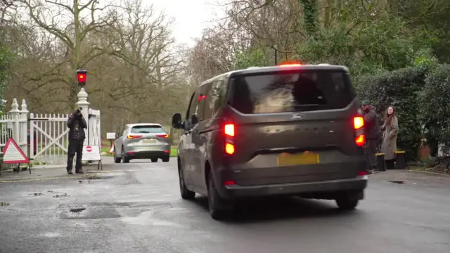 Two vehicles enter through the white gates of Royal Lodge