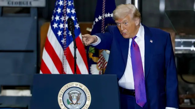 Donald Trump stands next to a podium and points to his right. An American flag is behind him