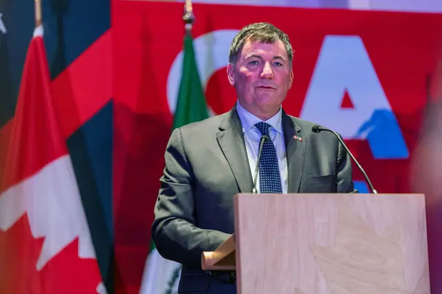 LeBlanc in green suit and blue dotted tie at an electern with a microphone before a red background and Canadian flag