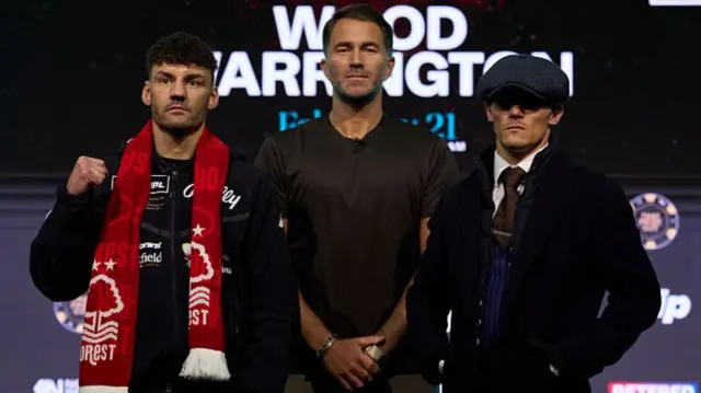 Leigh Wood holds up his fist and is wearing a red scarf around his neck, Eddie Hearn stands to the right with his arms down by his waist and Josh Warrington is further right wearing a flat cap and suit