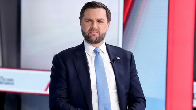 JD Vance sits at television station desk, wearing navy suit, white shirt and blue tie with his hands crossed