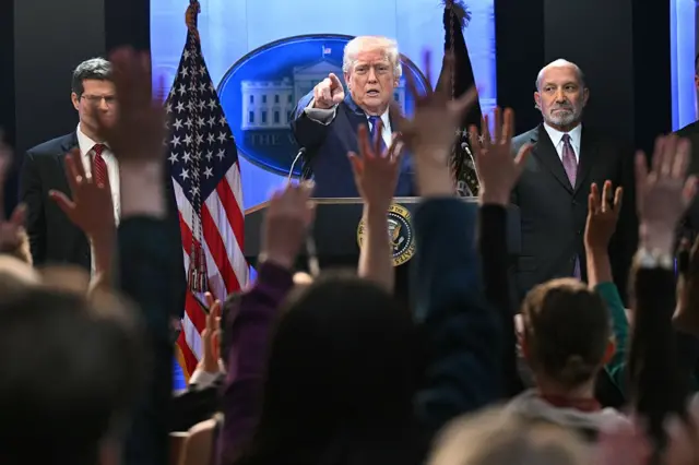 Members of the media raise their hands to ask questions to US President Donald Trump who is pointing at one of them.