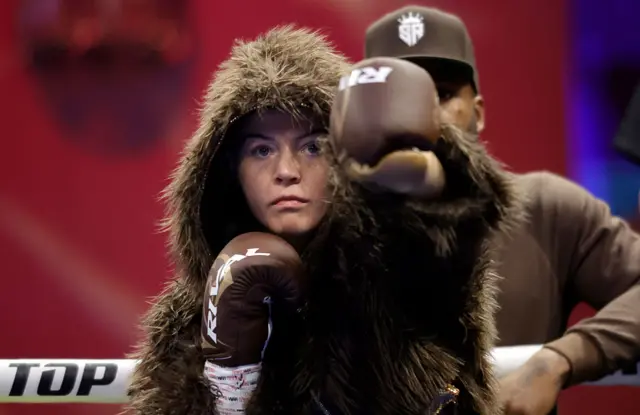 Sandy Ryan is wearing a fluffy brown jacket with a hood and is throwing a punch