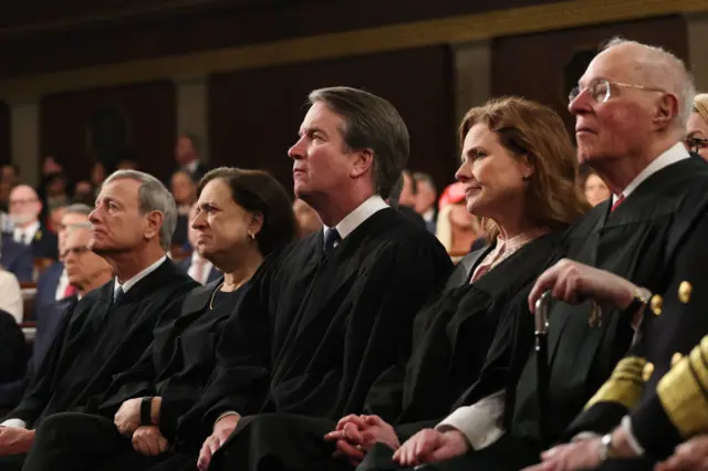 Supreme court justices sit next to one another in their block robes, looking up with their hands crossed