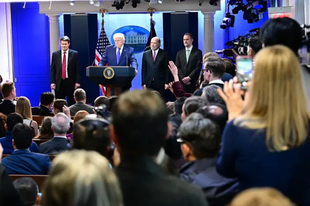 Long shot of the White House press briefing room showing the backs of reporters' heads and Trump standing at the podium with other officials