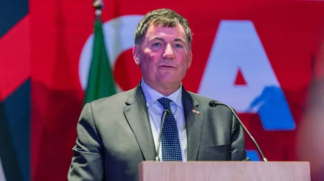 Dominic LeBlanc, Canada's US trade minister speaks from behind a podium. He is wearing a grey suit over a white shirt and a blue dotted tie