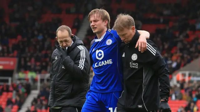 Victor Kristiansen (centre) holds his arm round the shoulder of a medic (right) as he is helped off the pitch