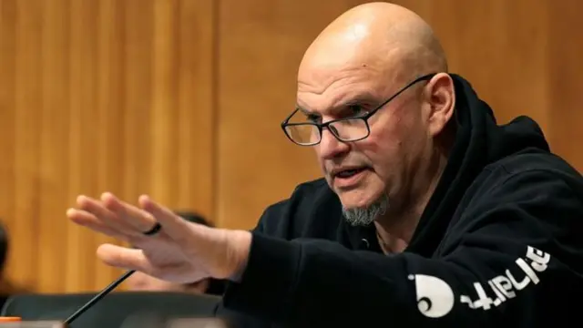 John Fetterman wears a carhartt jumper. he gestures with his hand ahead of him. he wears black glasses. he is sitting in a senate hearing.