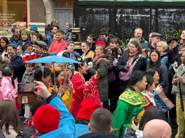 Crowd of people gathered in central london, surrounded by traditional flags and puppets to celebrate Lunar New Year