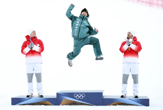 Men's giant slalom champion Lucas Pinheiro Braathen, from Brazil, jumps for joy on the podium alongside Swiss silver medallist Marco Odermatt and compatriot Loic Meillard, who won bronze, at Stelvio Alpine Skiing Centre