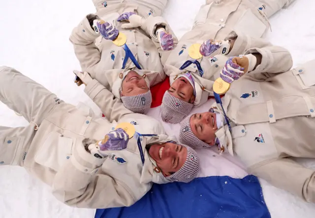 Four male athletes lay down on the snow on top of the French flag holding their gold medals