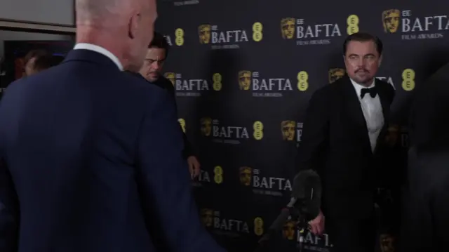 Leonardo DiCaprio dressed in a tuxedo looks at Colin Paterson, who is holding a microphone