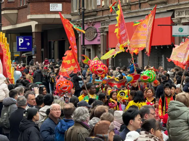 Crowd of people gathered in central london, surrounded by traditional flags and puppets to celebrate Lunar New Year