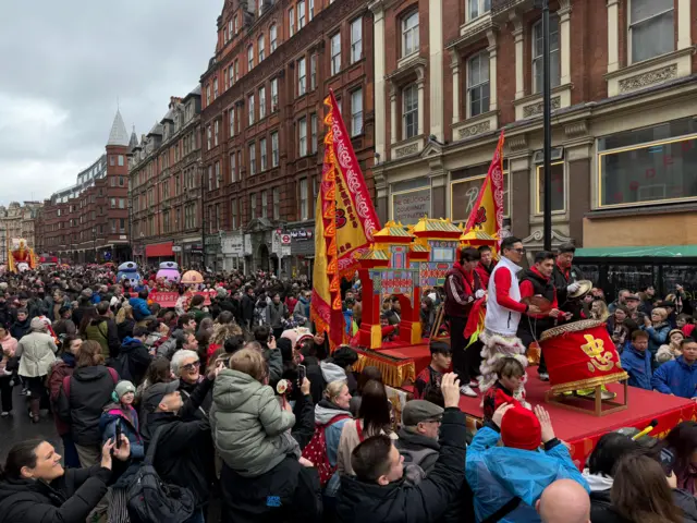 Crowd of people gathered in central london, surrounded by traditional flags and puppets to celebrate Lunar New Year