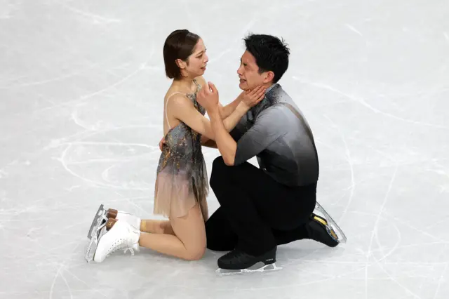 Two athletes kneel on the ice, the male is tearful and supported by his partner