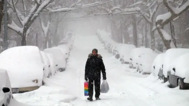 man walking on a snow-covered street next to cars submerged in snow